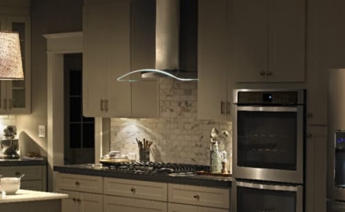 A wide view of a kitchen with a French door refrigerator, dual ovens and a wall-mount canopy hood over a cooktop